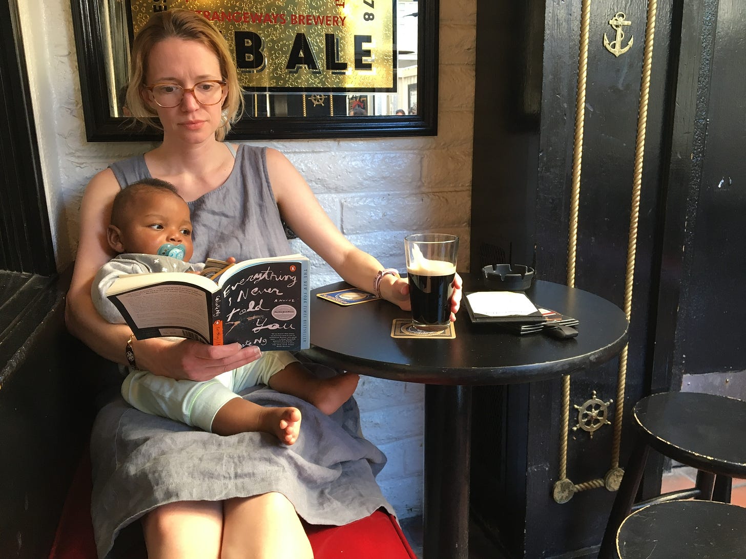 a white woman with a Black baby on her lap drinks a beer and reads a book at a bar