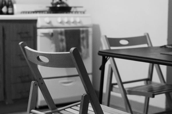 black and white photograph of two unoccupied wooden folding chairs at a folding table. behind the chairs and table (and out of focus) is a stove-oven with a tea kettle and a cast-iron skillet sitting on the burners