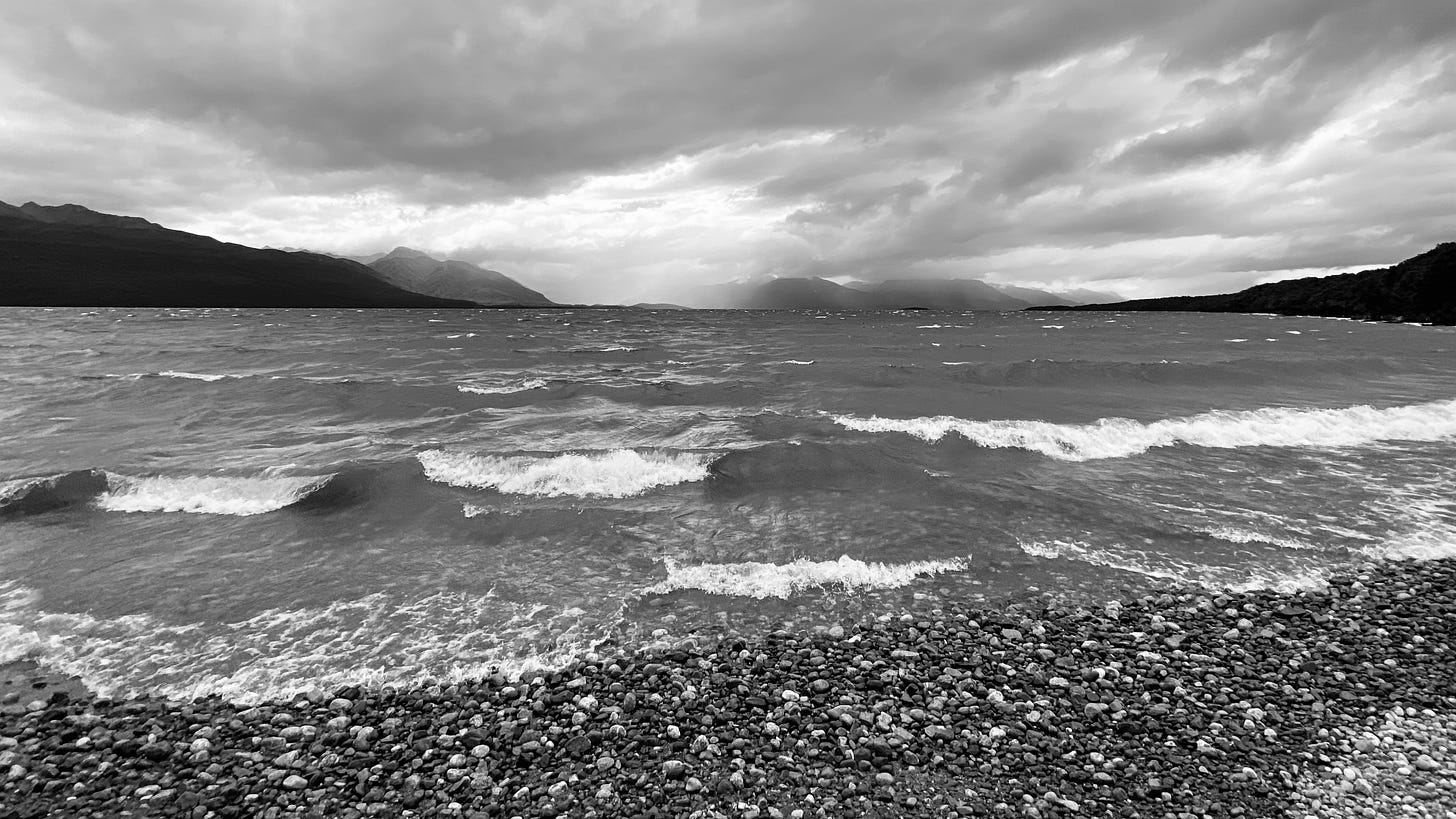 PerChatGPT: "A black and white image capturing a dynamic view of a pebbled lakeshore in the foreground, with small waves rolling in from the choppy water. The lake is surrounded by layered mountain ranges that fade into the distance under a dramatic, cloud-filled sky, suggesting a moody and windy atmosphere."