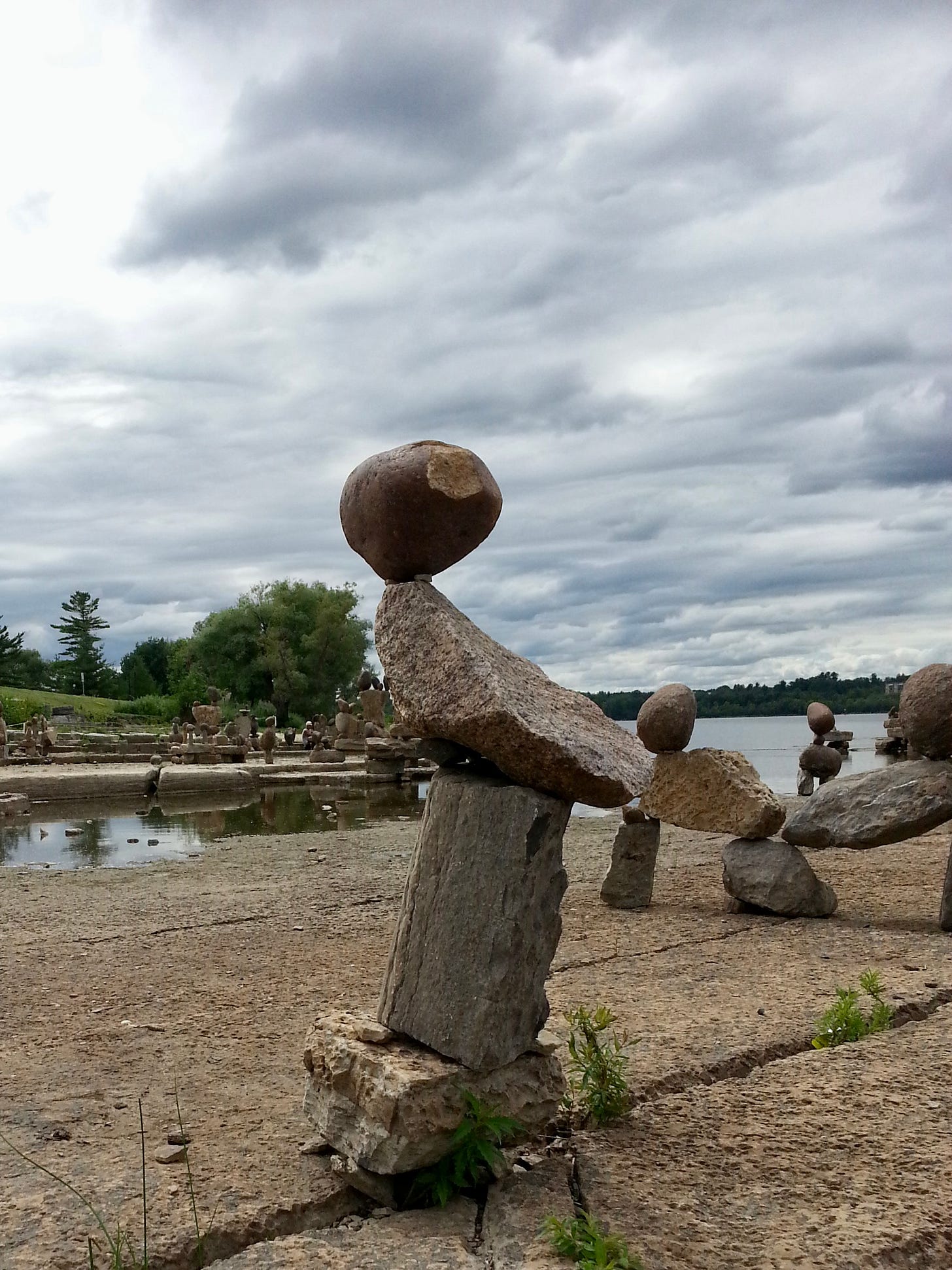 A stone inuksuit stands in the foreground of a flat stone ledge on the edge of a body of water, under a cloudy sky and with a treelined shore in the background.