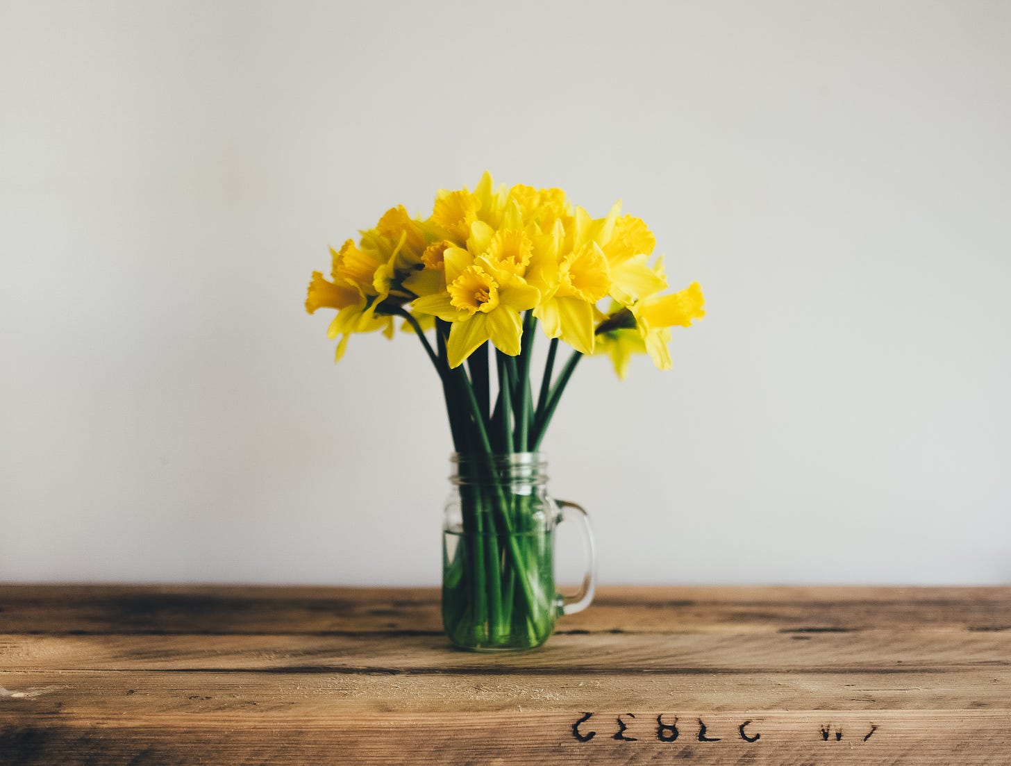Yellow flowers with green stems sit in a clear glass mug serving as a vase. The table it sits on is made of brown wood. The wall behind the flowers is white.