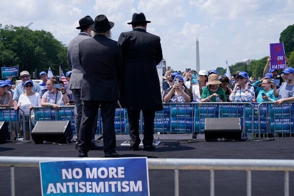 A sign that reads “no more antisemitism” hangs on a barricade in front of a stage in Washington, D.C. Two men in black suits and one man in a gray suit, all wearing hats, face the crowd, with the Washington Monument in the distance.