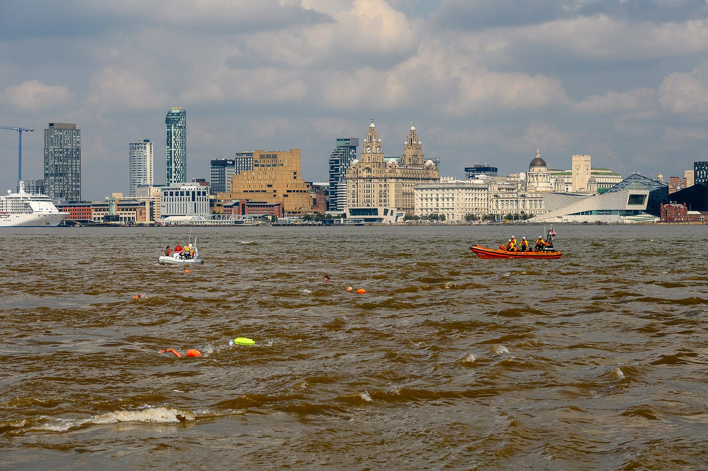 People with toe floats swimming across the River Mersey. There are safety boats in the area and the skyline in the distance.