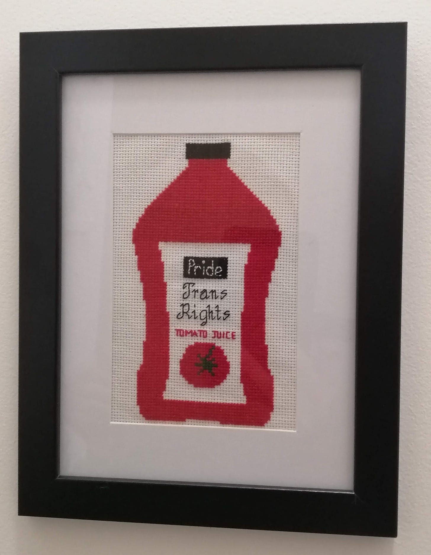 Cross stitch of what looks like a Keri brand tomato juice bottle but the brand is Pride and it says trans rights tomato juice. In a black frame with white mat.