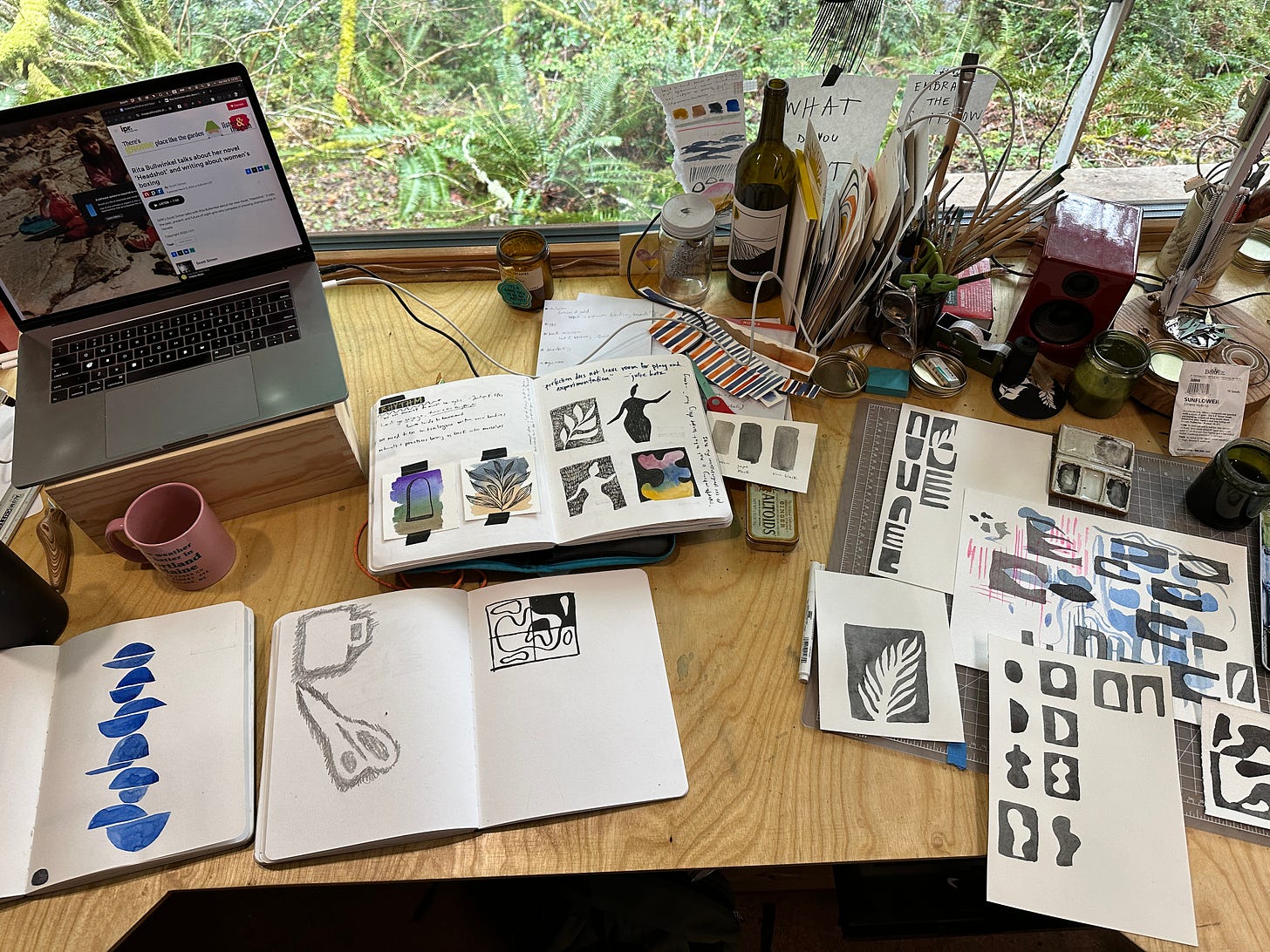 A photo looking down at a desktop covered in notebooks with drawings and paintings and a computer and cup of coffee