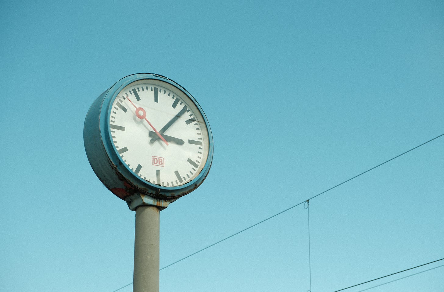 Poste relógio antigo na estação central de trem de Dusseldorf, Alemanha. O relógio é redondo e azul, ponteiros pretos e vermelho. Possui sinais de ferrugem. O céu é azul claro e há fios de transmissão na diagonal inferior direita.