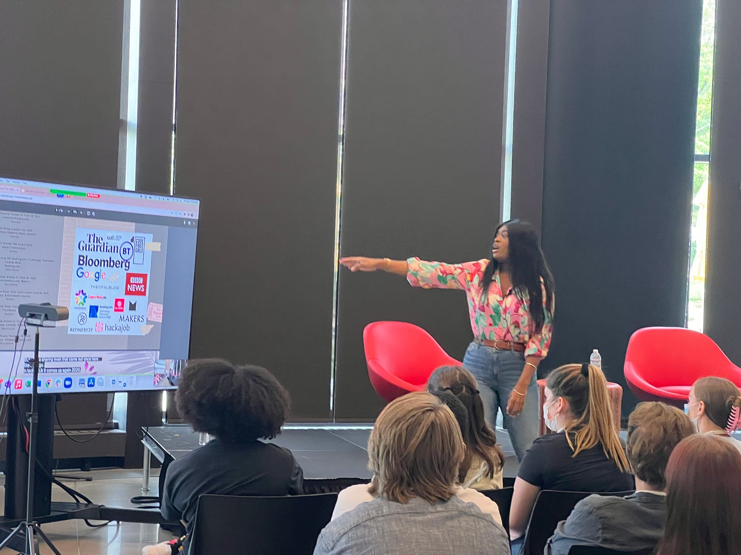 A black woman with pink print button up collared shirt rolled up her sleeve. She is pairing that with high waist blue denim jeans and two gold bangles. She is facing a screen where her slides are being shown & has waist length straight black hair.