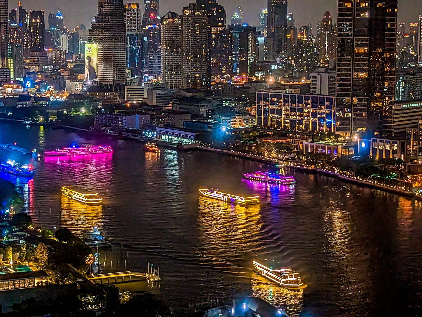 Boats lit up in yellow, blue, and purple drift along the Chao Phraya River, the glittering Bangkok skyline behind. 