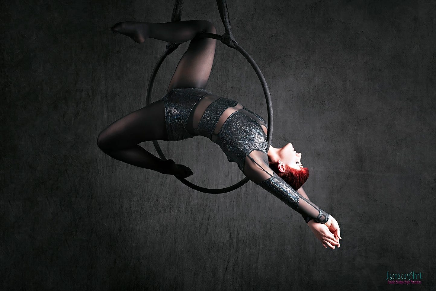 nikkita nouveau, a person wtih white skin and bright red hair, hangs sideways from a lyra hoop. their arms are crossed behind their head below, and their knees are bent to hold themselves on the hood. they wear a black and mesh bodysuit against a black background. the artist attribution in the bottom right corner says "jenu art"
