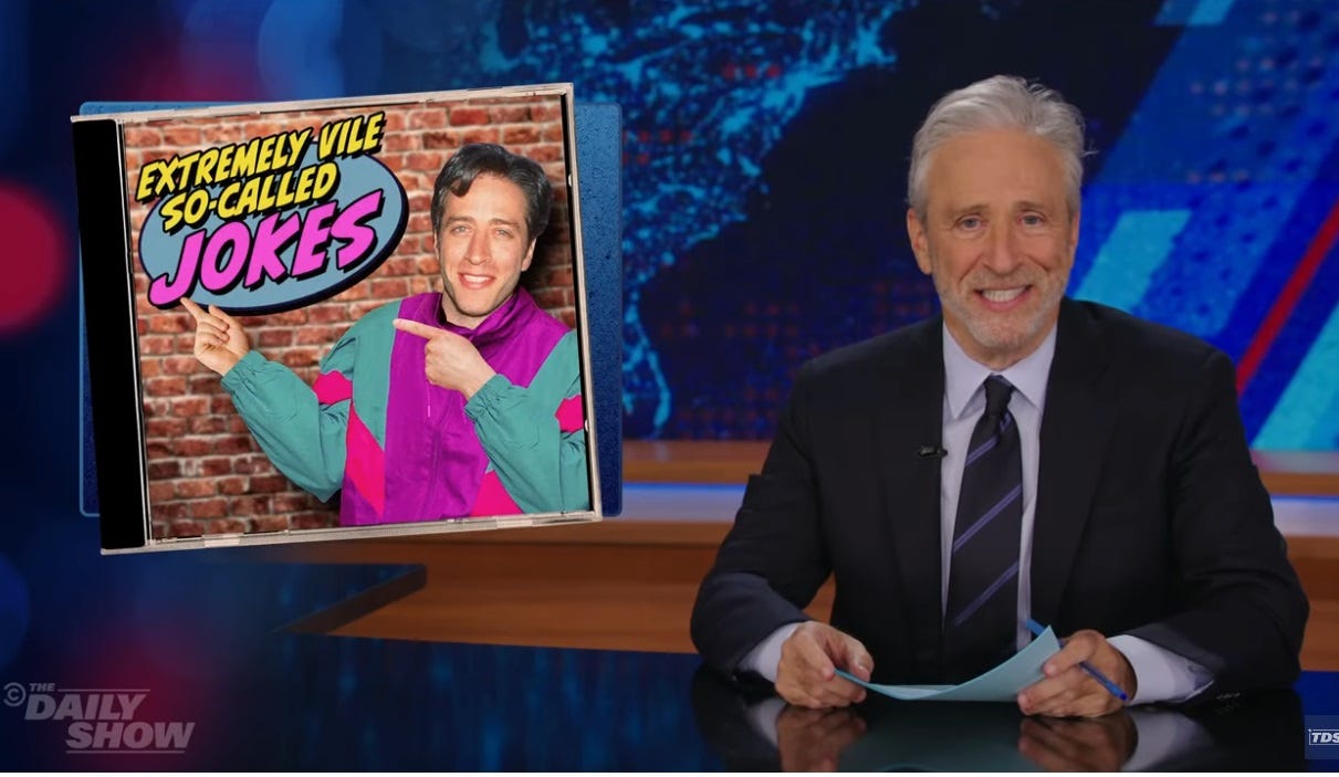 Video screenshot of Jon Stewart, smiling, at the Daily Show desk, with a graphic of a fake CD titled "Extremely Vile So-Calles Jokes,' depicting Stewart as he looked 30 years ago