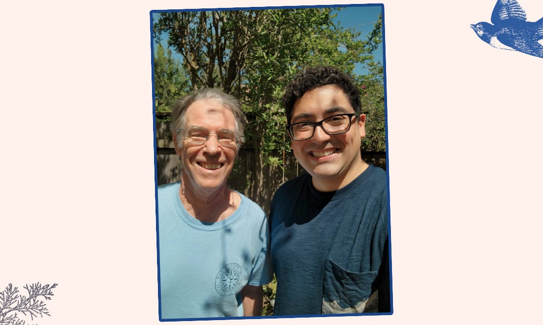 Dois homens sorrindo para a foto e usam óculos. O mais velho tem cabelo branco e camiseta azul. O segundo, cabelos encaracolados pretos e camiseta azul escura. O fundo tem árvore e sol.