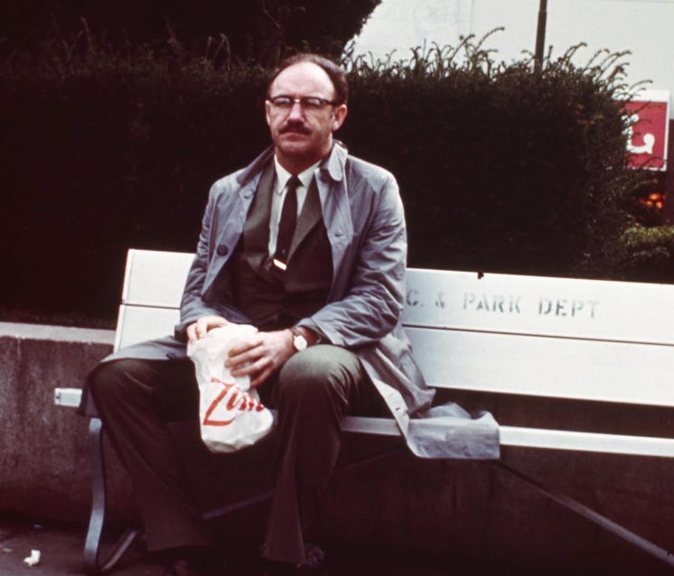 The actor Gene Hackman, a balding man dressed in a tranchcoat over a brown suit, white shirt, and black tie, slumps forlornly on a park bench, holding a white take-out bag from a diner on his lap