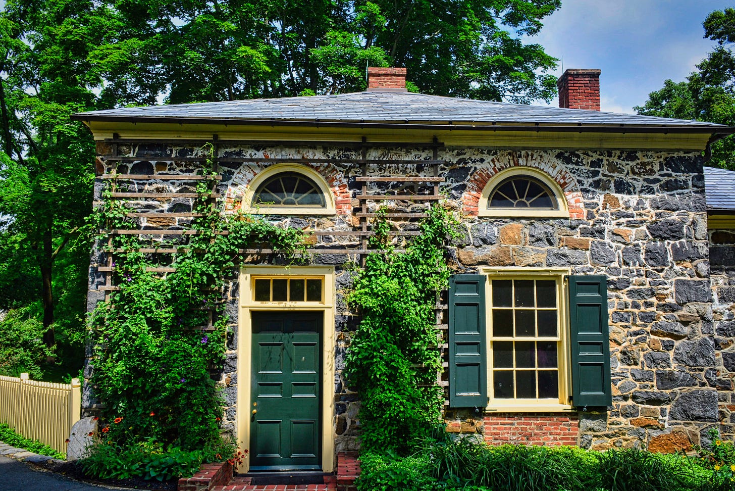 A stone house with a hip roof and chimneys showing, a dark green front door, and a shuttered window with yellow trim; a yellow picket fence is on the left and shrubs climb around the entrance