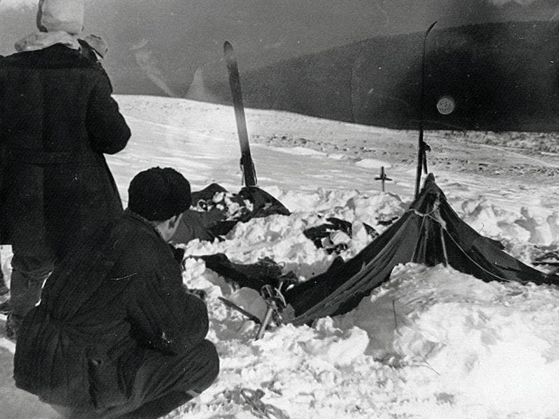 Black and white image showing a barren, snow-covered field. There are the destroyed ruins of a campsite there: a torn up tent and disturbed snow. Two men are nearby, presumably part of the team that found it and took the photo.