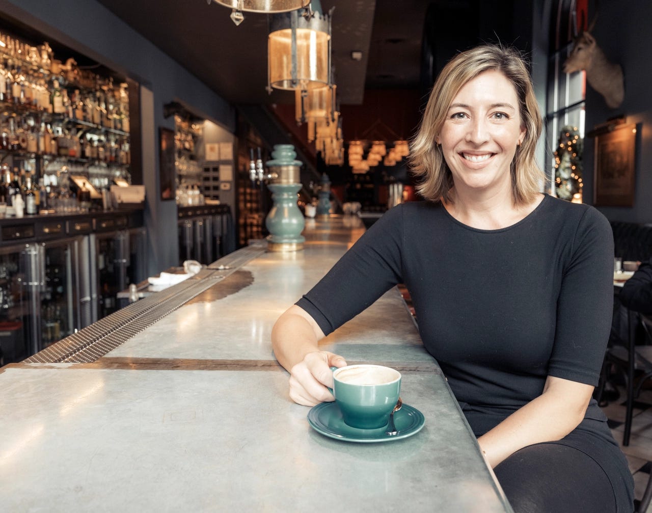 Photograph of Molly Wood sitting on a high stool at a bar with a cup of coffee. She is looking at the camera with her right hand on the handle of the cup which has a saucer and a spoon on the saucer.