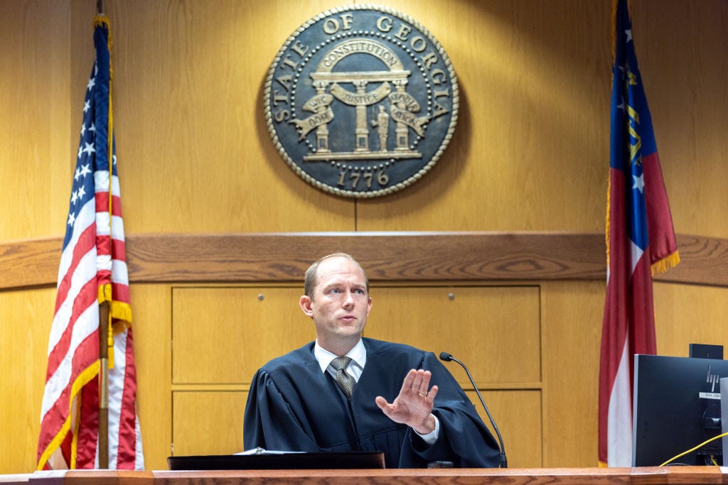 A clean shaven white main in a silver tie and a judge's black robe gestures with his left hand, sitting between US and Georgia flags. Above his head, on a wood paneled wall, is the seal of the state of Georgia.