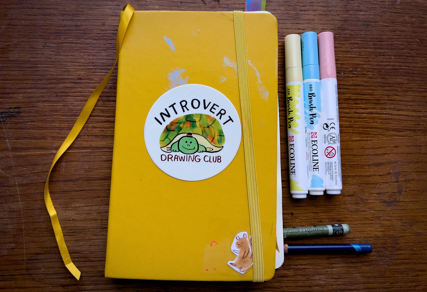 sketchbook with introvert drawing club sticker on cover alongside markers and a wax crayon