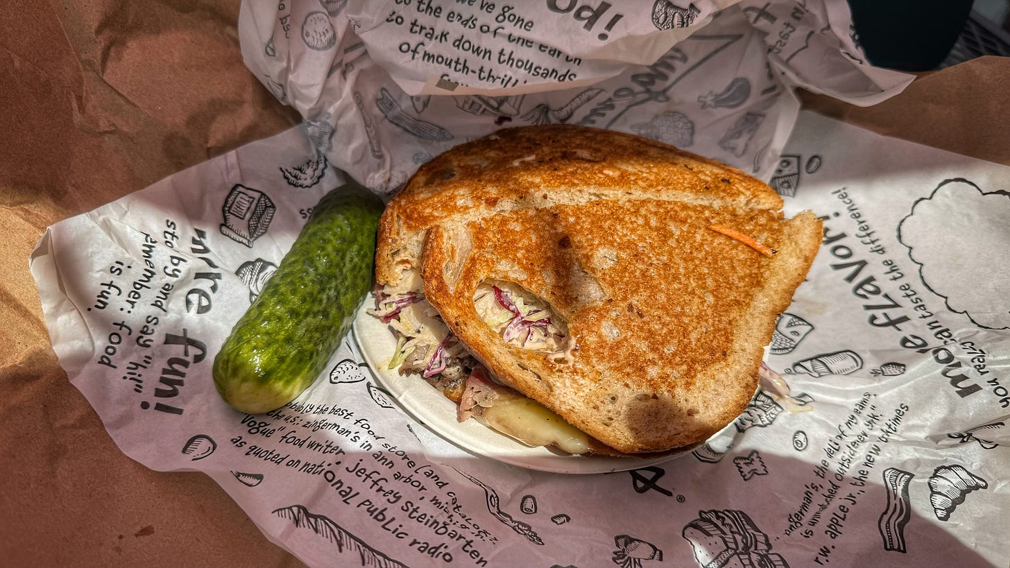 A Jenny's Fix sandwich on a paper plate, beside a half-sour Kosher pickle. It's all sitting on a piece of Zingerman's wrapping paper, with hand drawn illustrations of products and quotes from guests.