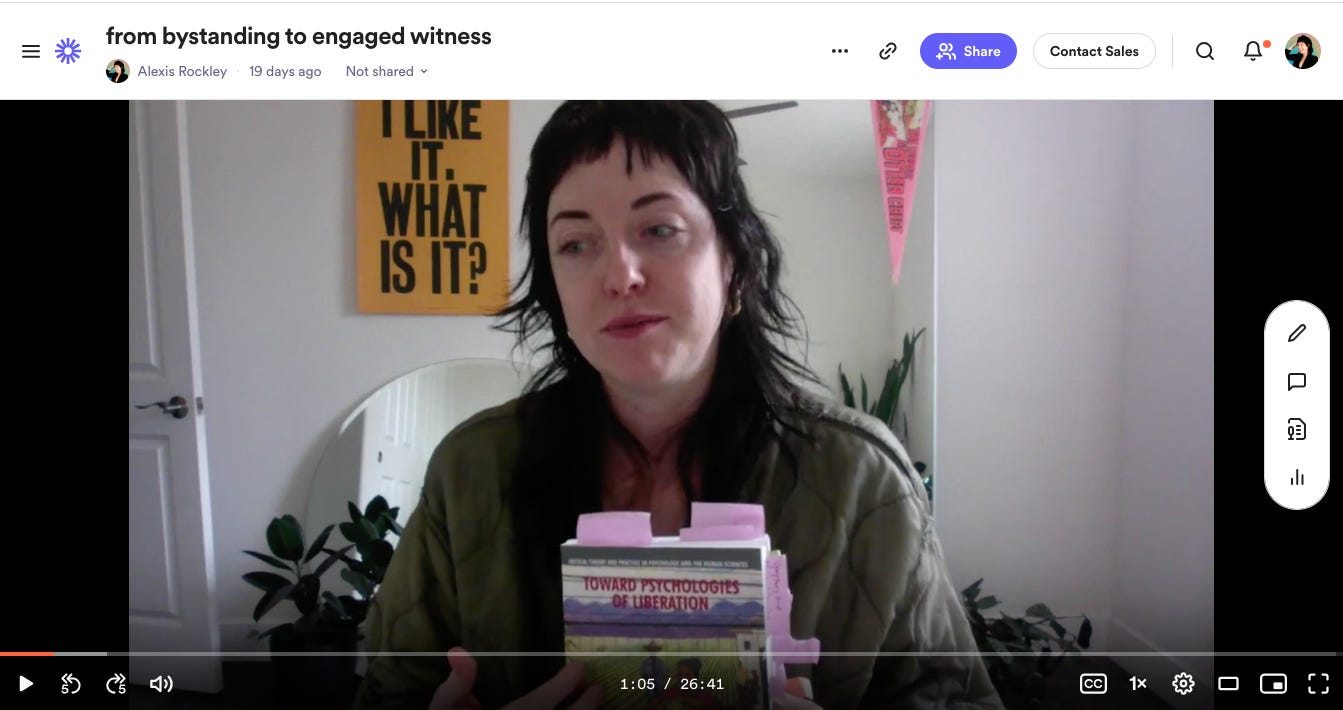 a screenshot of Alexis' video paused at 1-minute in, labeled "from bystandng to engaged witness." She is holding up a copy of the book Toward Psychologies of Liberation, looking slightly offscreen, wearing a green jacket and with layered brown hair. In the background are plants, two mirrors, and a poster that says "I like it. What is it?"