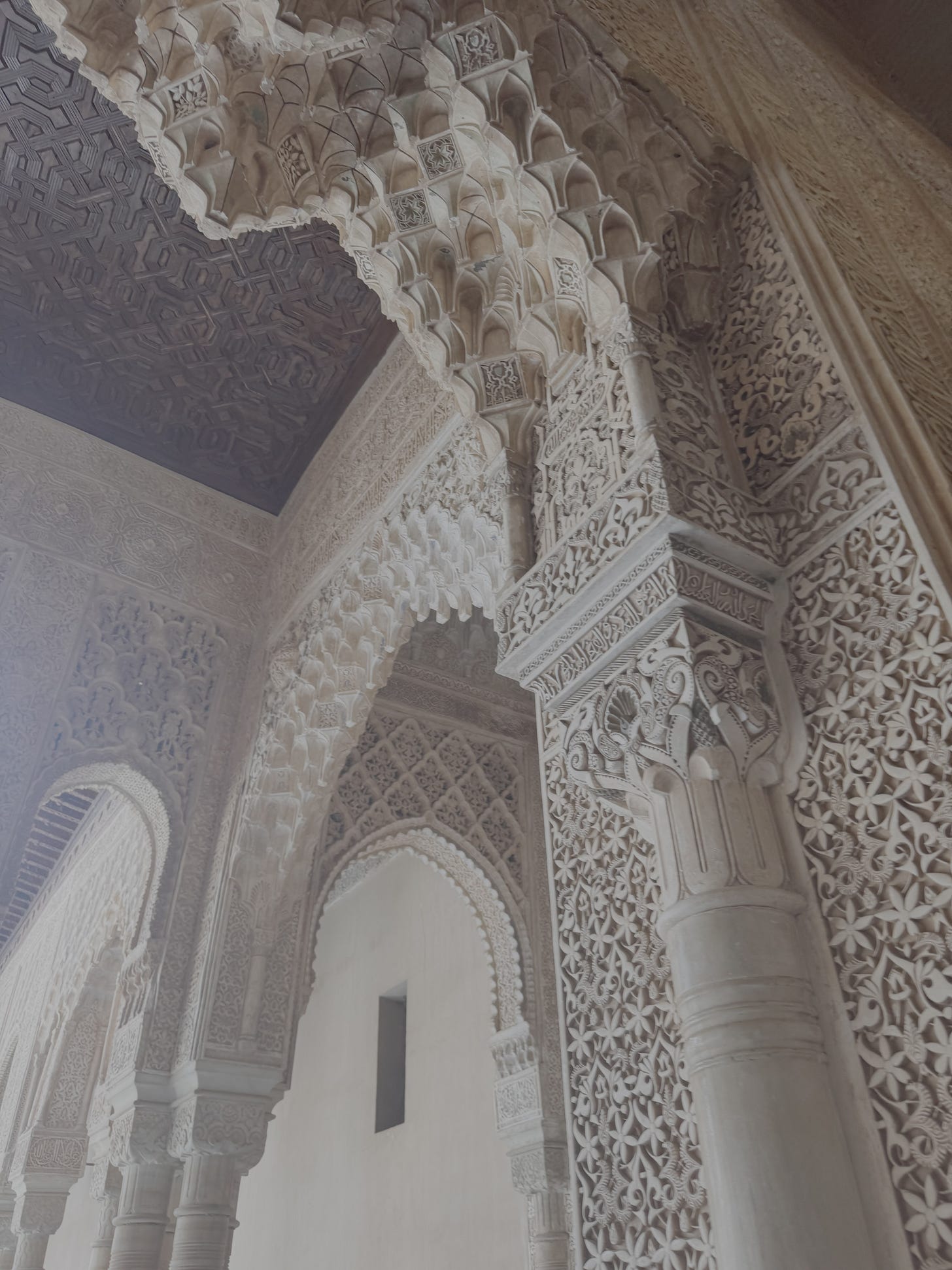 Ornate stone arches and columns at the Alhambra in Granada, Spain, showcasing intricate Islamic geometric and floral carvings beneath a dark, patterned wooden ceiling.