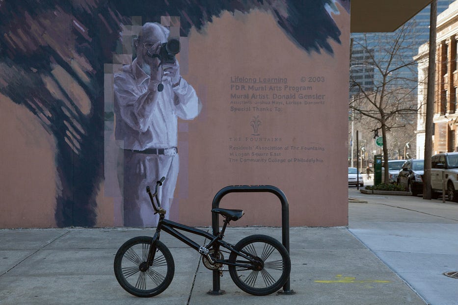 Mural of a photographer. Bike rack is shaped like a bike in the foreground, making it appear that the man on the wall is photographing the bike.