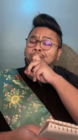 A man with glasses wearing an ornate sash thinks deeply, tapping a pen to his lips and taking notes. Over-exaggerated intellectual reaction for comedic flair.