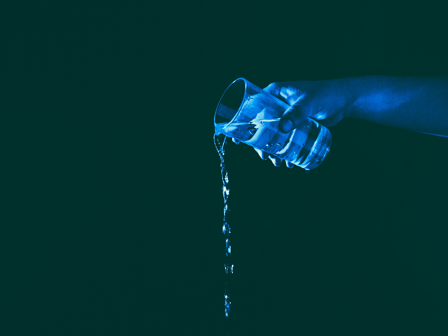 A blue hand pouring water out of a clear glass over a black background. Light refracts and the hand is illuminated by the reflections in the water.