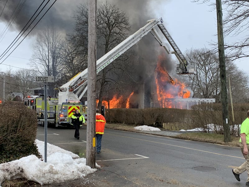 A large fire truck with a jointed ladder and a platform with a water cannon extends the ladder from its position on the road and above a hedge to a position near a house totally engulfed in flames. 