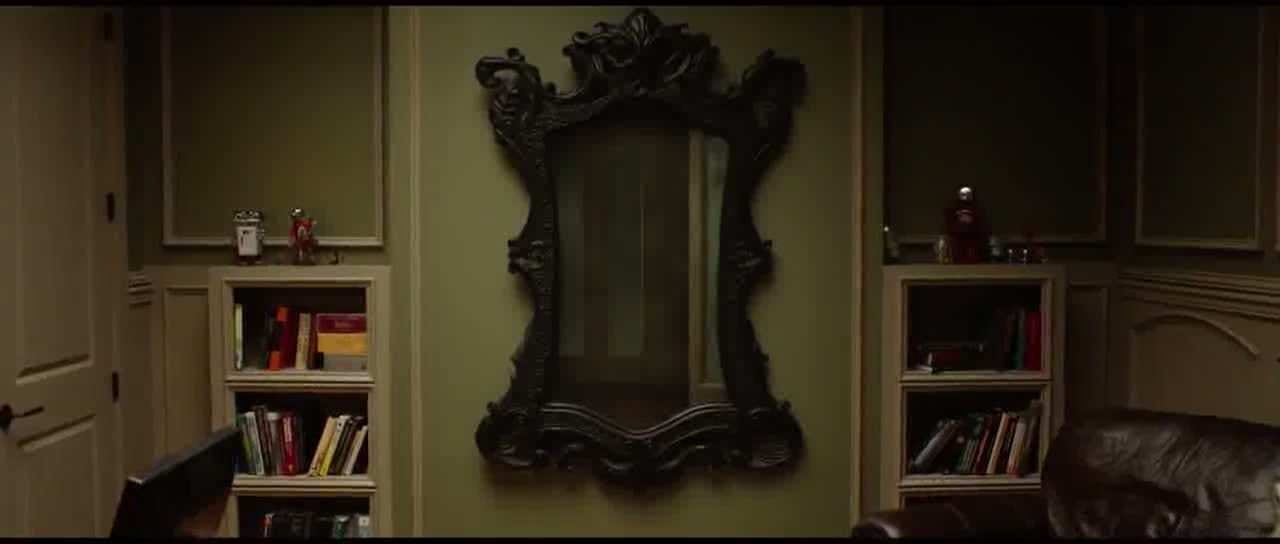 Scena del film Oculus in cui c'è un grosso specchio antico con la cornice scura in una stanza dalle pareti verde chiaro, due piccole librerie bianche con dei libri e un divano scuro