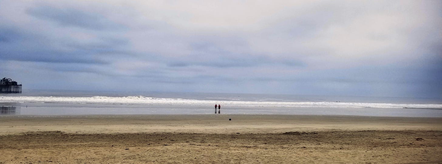A stretch of sand in the foreground. In the distance, two tiny silhouettes walk toward the breaking ocean surf, showing the immensity of the ocean.