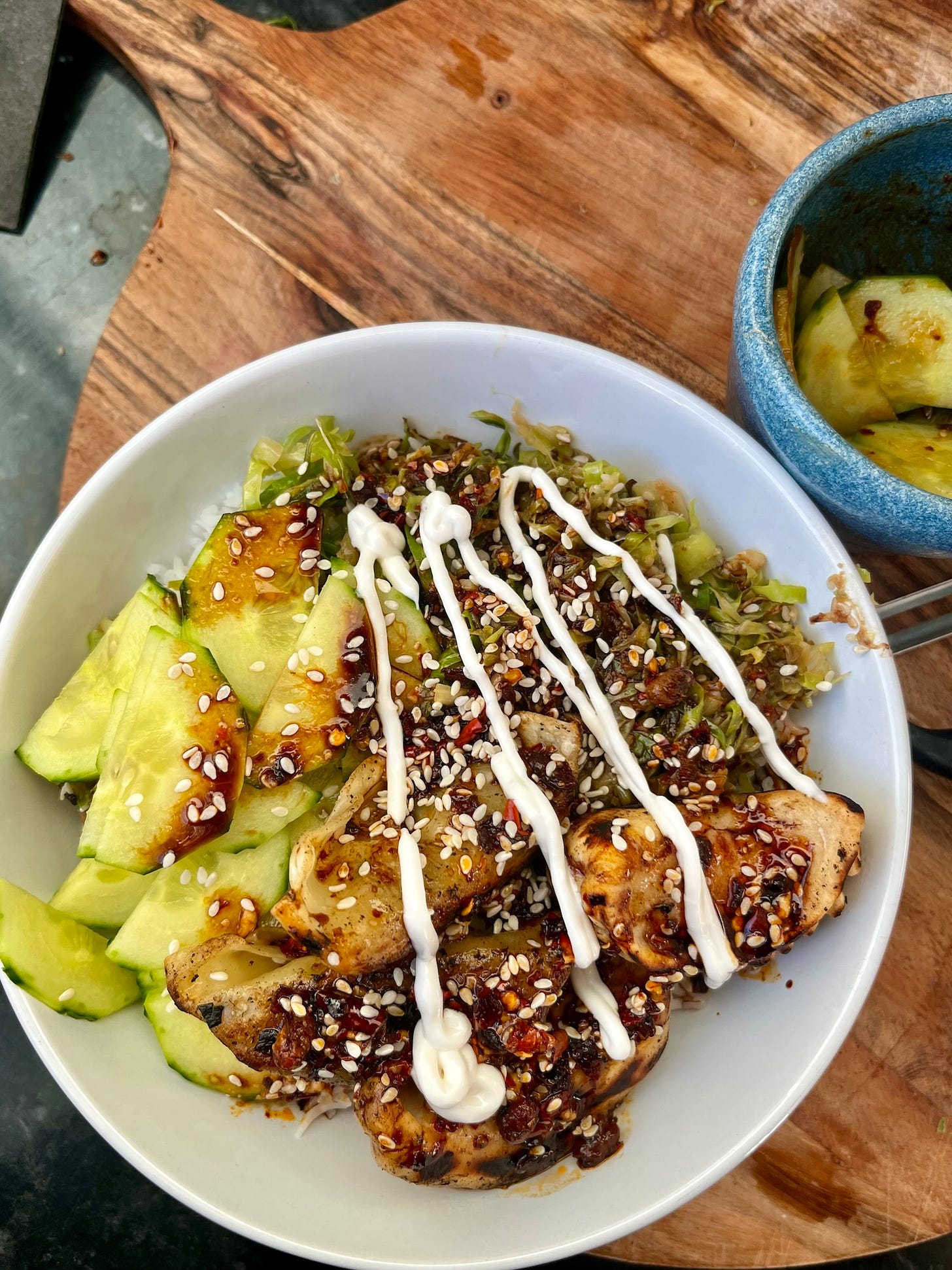 a white bowl containing rice, stir fried cabbage, cucumber, dumplings and sauce, sesame seeds and mayo next to a smaller bowl with cucumbers all on top of a wooden cutting board