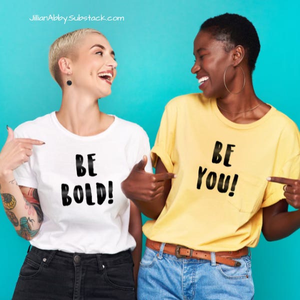 A white woman with short blonde hair and a black woman with short black hair are pointing at messages on their t-shirts. The blonde woman's white t-shirt says Be Bold and the other woman's shirt is yellow and says Be You.