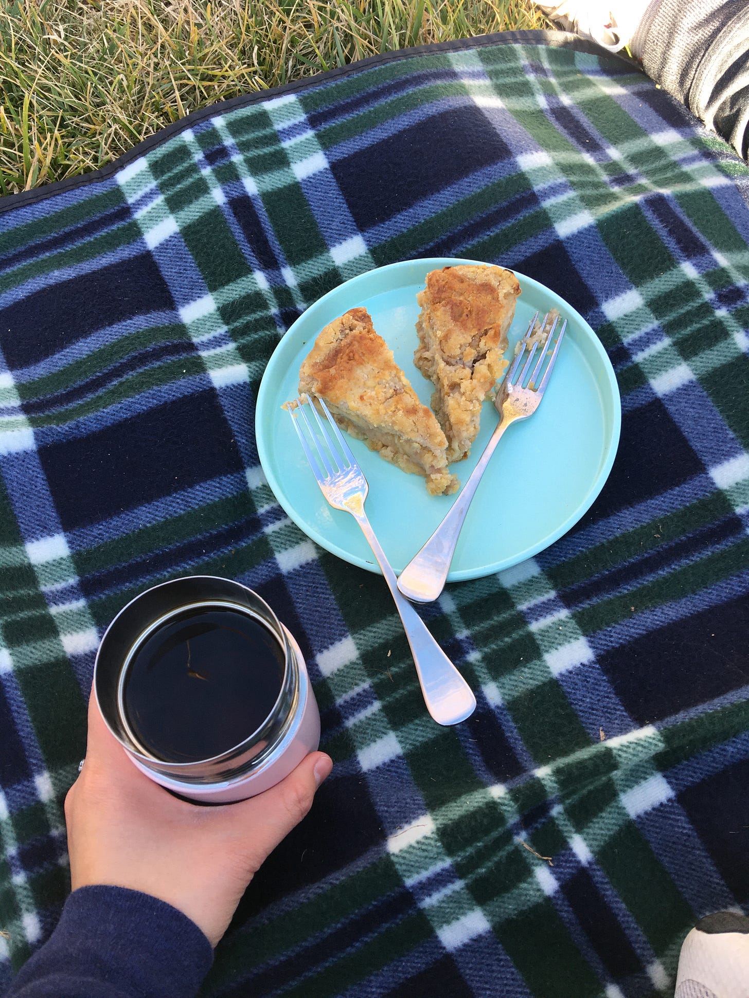 Two slices of apple crumble cake on a picnic rug with a reusable coffee cup filled with filter coffee.