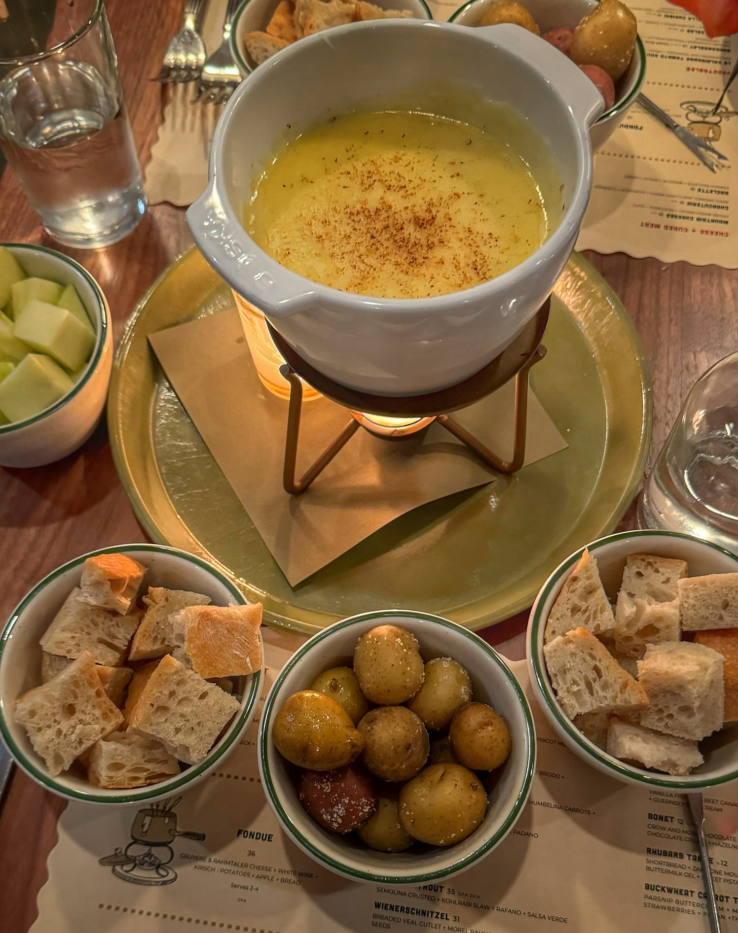 A small fondue pot full of melted cheese, being warmed by a tealight. Surrounding the fondue pot are small bowls with chunks of bread and apple and baby potatoes with large flakes of salt on them. 