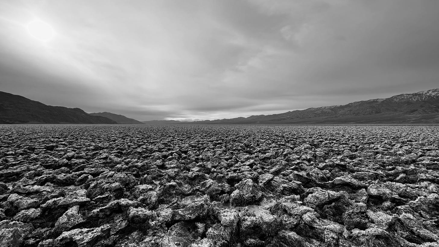 PerChatGPT: "A black and white photo displaying a vast field of jagged, cracked earth stretching towards a mountain range in the distance. The rugged terrain dominates the foreground, leading to the mountains shrouded in haze. The sky is overcast with the sun peeking through the clouds, casting a soft glow over the scene."