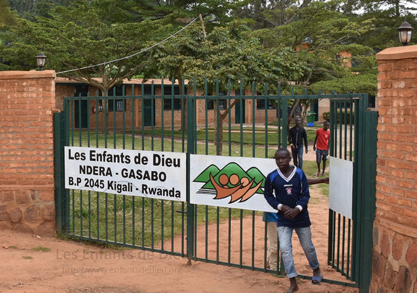 Rwandan boy, age about 10, welcoming us to the Les Enfants de Dieu home. He is standing at the front gate, with the door open and grounds and buildings in background