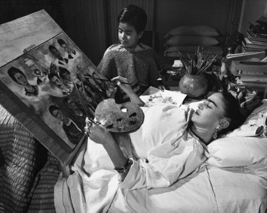 Black and white photo of Frida Khalo painting, laying in bed with an easel designed for her to paint in bed