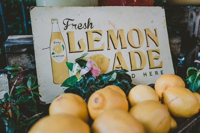 Several lemons sitting on a tray in front of an old-fashioned metal sign that reads “Fresh Lemonade Sold Here.”