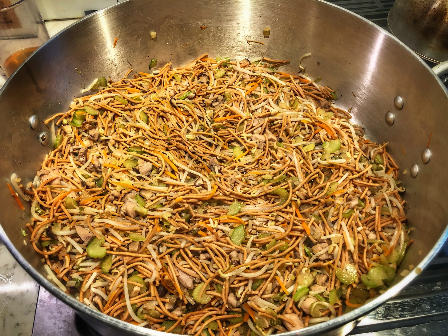 A very large pan that covers several burners on a stove, holding finished chow mein. Noodles, and small morsels of onion, celery, carrot, bean sprouts, and chicken are visible. 