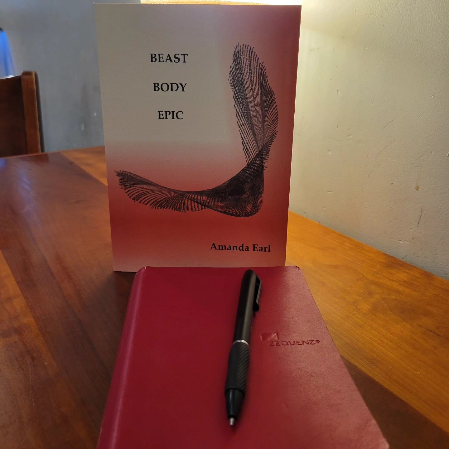 Book on a table with a red journal and a pen, background lighting.  TEXT on red and white book cover: Beast Body Epic/Amanda Earl; visual poem in centre of cover.