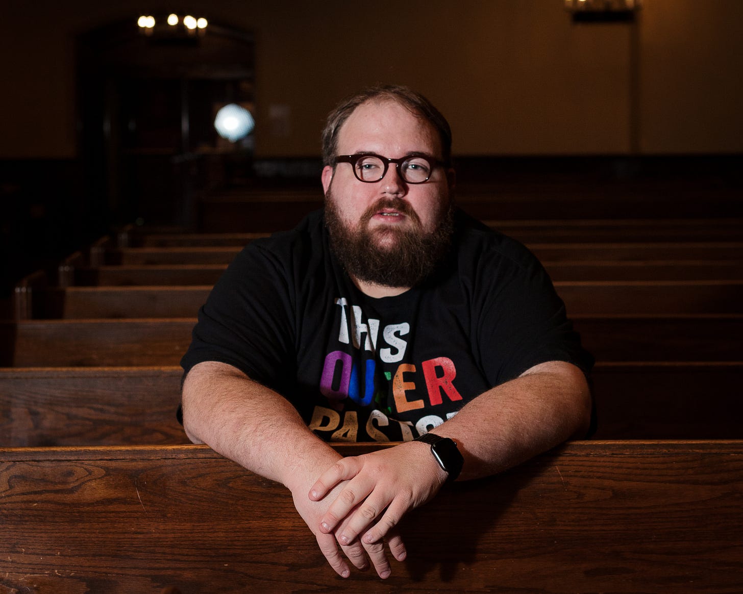 Photo of Ben Huelskamp sitting in a pew wearing a shirt that reads "This Queer Pastor Loves You."