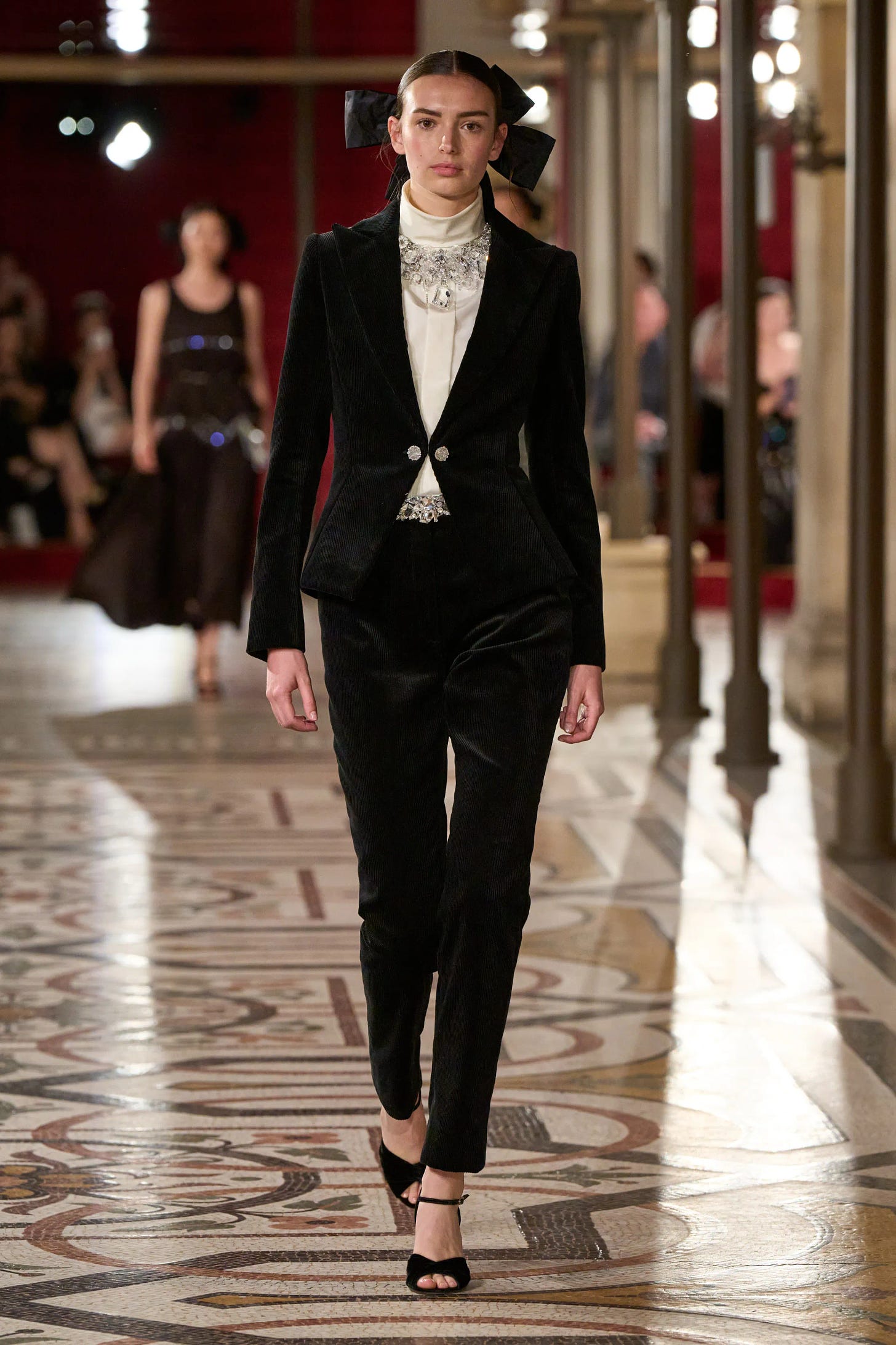 A light-skinned model wears a tailored black velvet suit that has a nipped waist and slim trousers. Beneath, she wears a high-collared cream satin blouse overlaid with a crystal necklace.