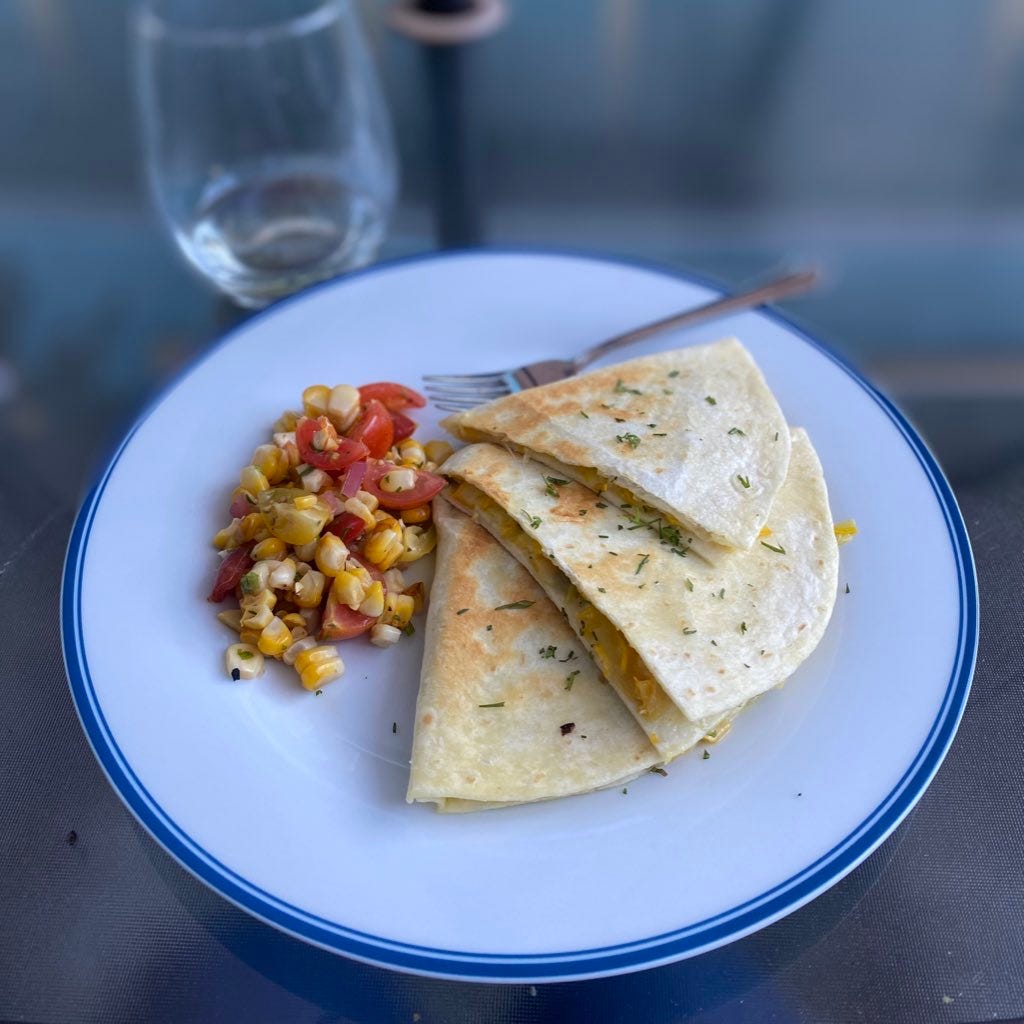A large white plate with a blue rim holding three triangles of browned quesadilla, the filling of cheese and zucchini just visible at the front edges. Next to the arrangement of quesadilla is a helping of the corn and tomato salsa described above. Cilantro is sprinkled over everything, and a glass of white wine is visible but blurred in the background.