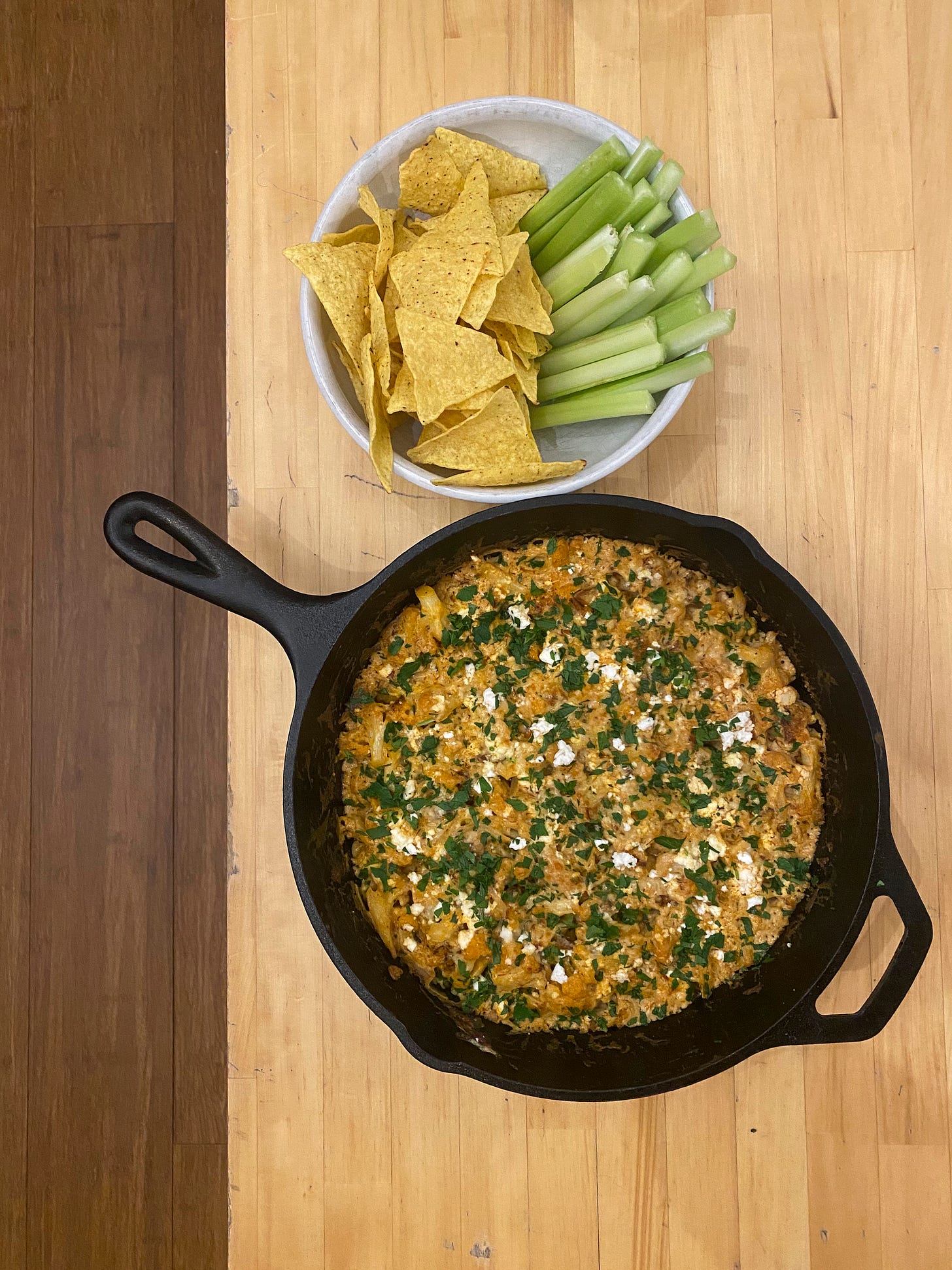 From above, on a light wood coffee table, a cast iron pan of the dip described above, a burnt orange colour made creamy with cheese. On top are pieces of feta and sprinkles of parsley. Above it is a white bowl with tortilla chips on one side and celery sticks on the other.