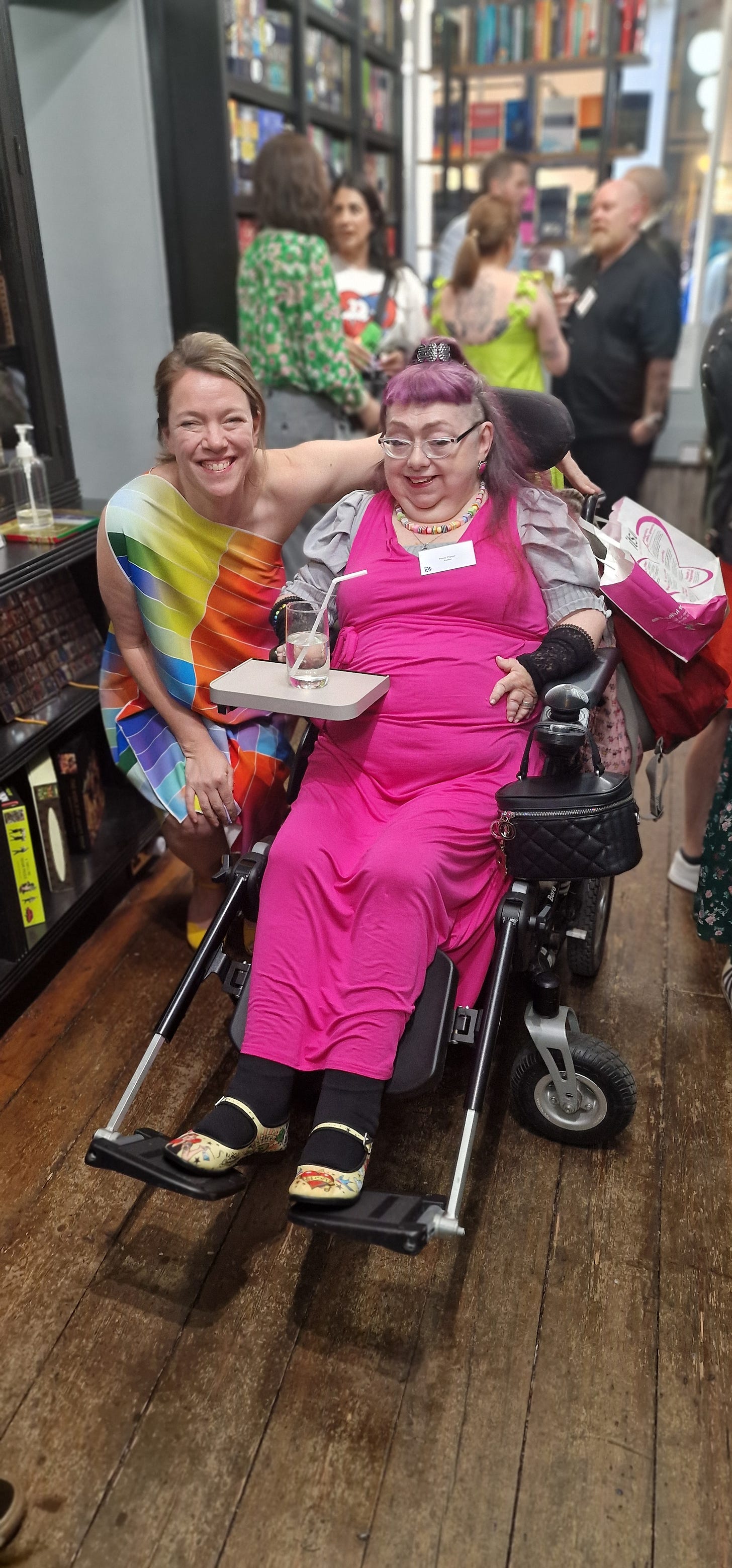 Penny is dressed in a bright deep pink dress and a grey under top. She has a short blunt vintage style fringe and longhair part of a metal her class. She has pale white skin and wears transparent cats eyes  glasses. She is sitting in her wheelchair which has a small grow table attached, which is a drink. Besides her is her agent in a striped multicoloured dress, crouched down to be level with Penny. Both of them are smiling, in a old bookshop, at a literary event in central London.