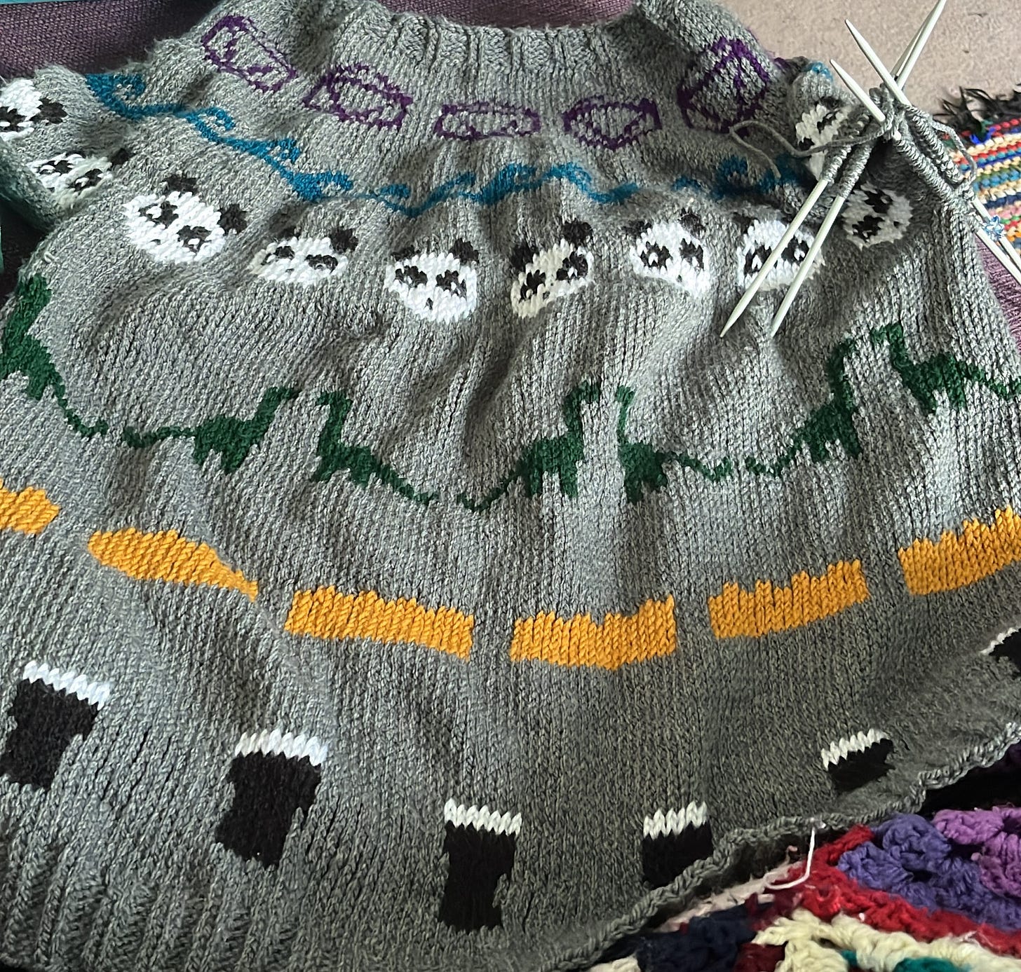A top down knitted jumper in grey with rows of repeated motifs: A purple D20, blue wavs, a pandas face, dinosaurs, yellow lego brick and a pint of dark beer
