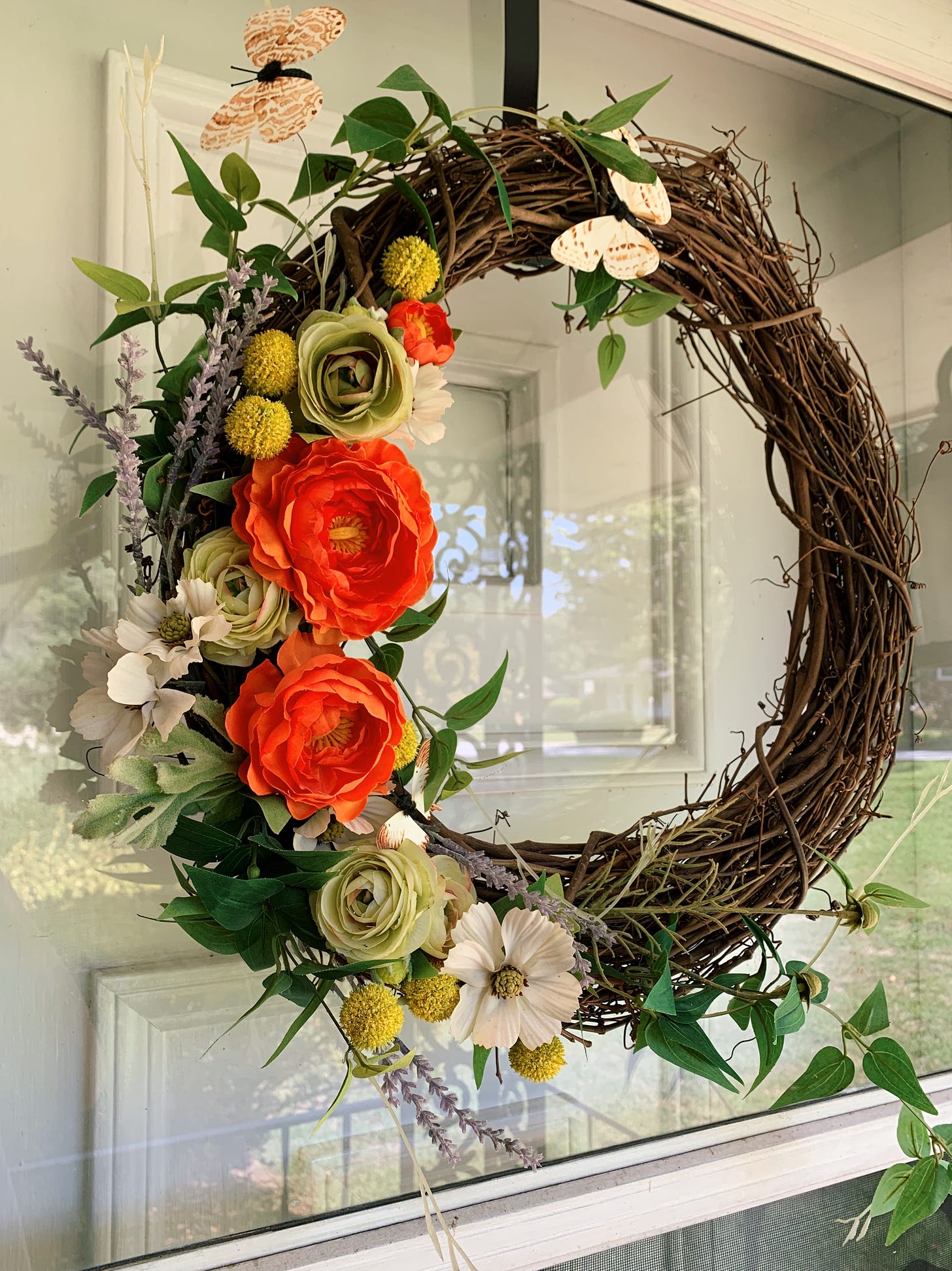 A handmade wreath hanging from a glass storm door. The wreath is made of brown branches woven together. Half of the wreath is covered in fake flowers with a palette of orangey-red, yellow, white, and lavender. There are three paper-like butterflies on the wreath.