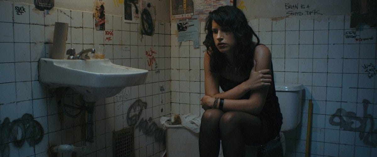 In a disgusting, graffitied and flyered dirty white tiled bathroom, a tall olive complexed woman with long dark wavy hair is sitting on the toilet with her long arms resting on her knees as she leans forward looking forlorn.