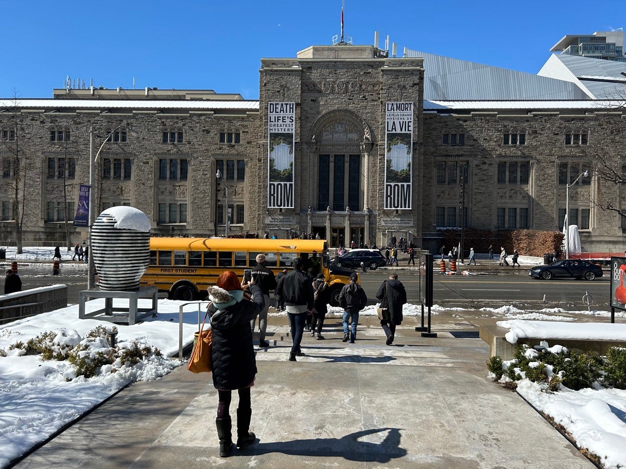 A large yellow bus parked on the street, with people walking on stairs towards the bus. There is a large gothic style building in the background