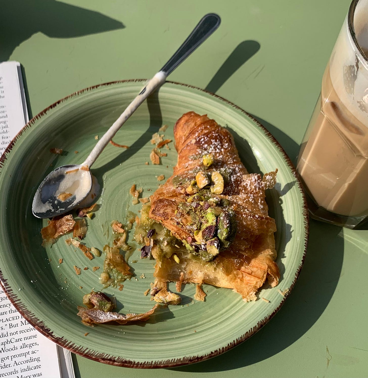 My favorite pistachio croissant on a green plate set on a green table with a partial view of iced coffee in the side of the photo